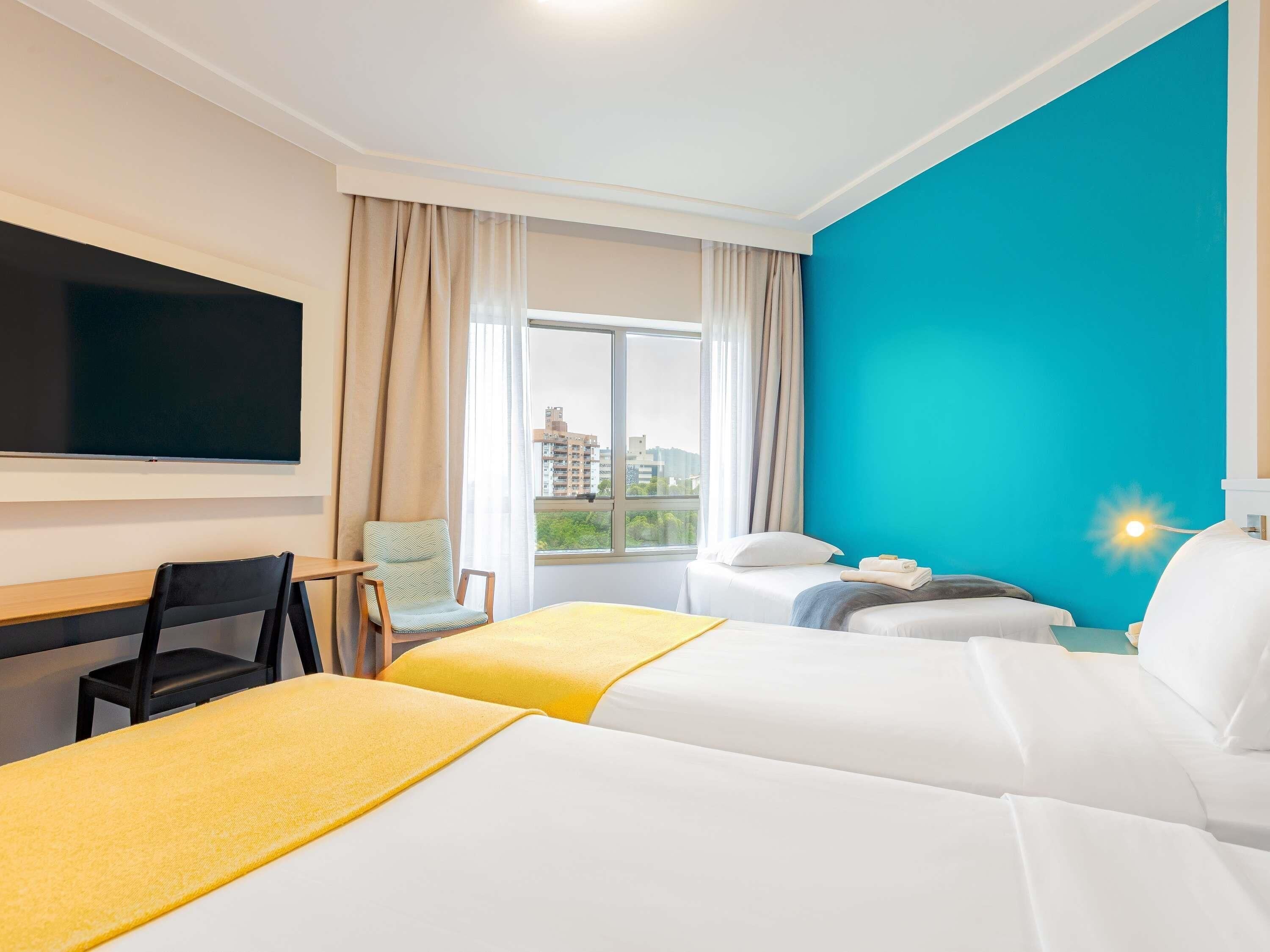 Quarto de hotel com duas camas, uma escrivaninha, uma cadeira, uma televisão e uma janela com cortinas.