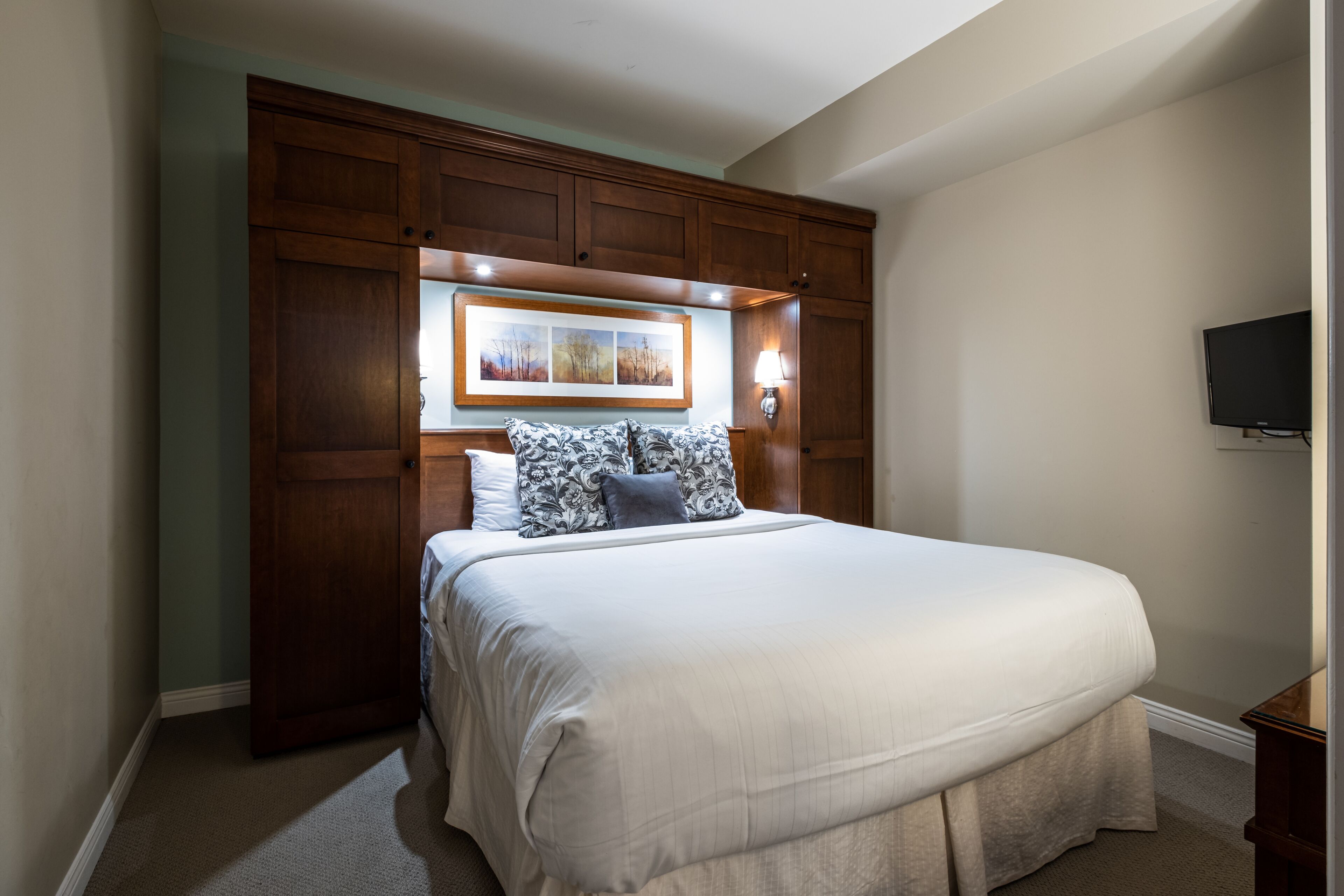 A neatly made bed with a white comforter and patterned pillows, a wooden headboard with a framed picture, and a wall-mounted TV.