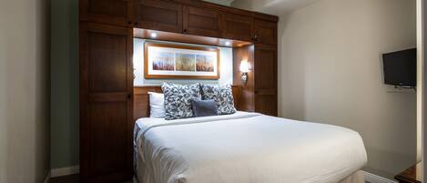 A neatly made bed with a white comforter and patterned pillows, a wooden headboard with a framed picture, and a wall-mounted TV.
