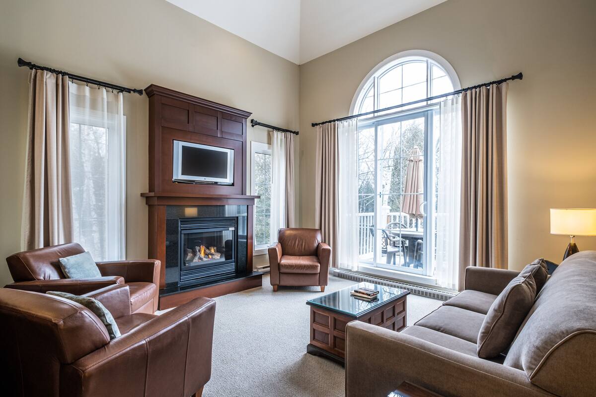 A living room with leather sofas, a fireplace, a flat-screen TV, and large windows with curtains.