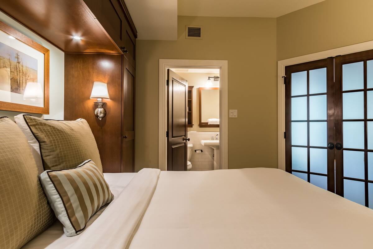 A hotel room with a bed, a nightstand, a lamp, a door leading to a bathroom, and a window with frosted glass.