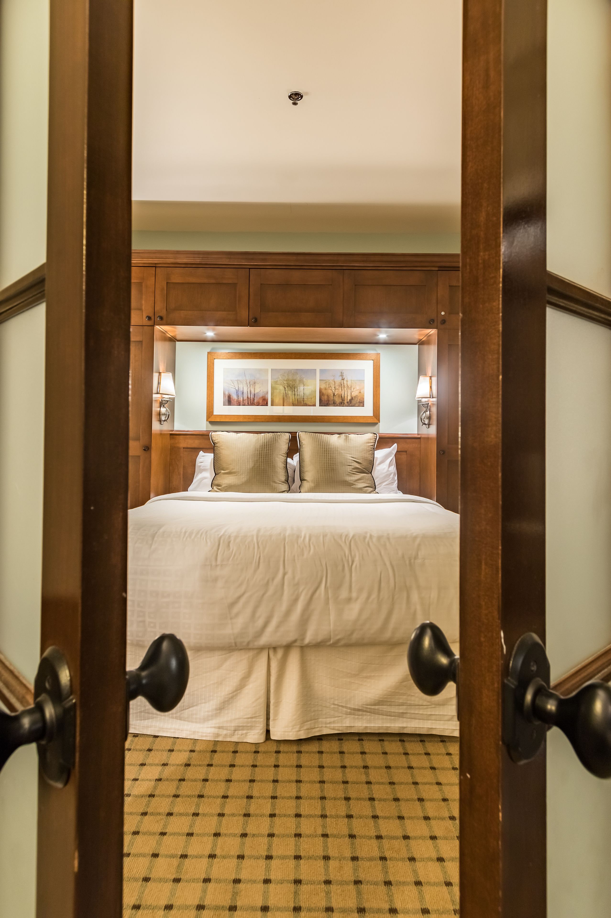 Una habitación de hotel con cabecera de madera, una cama grande con ropa blanca, dos lámparas de noche y un cuadro enmarcado sobre la cama.