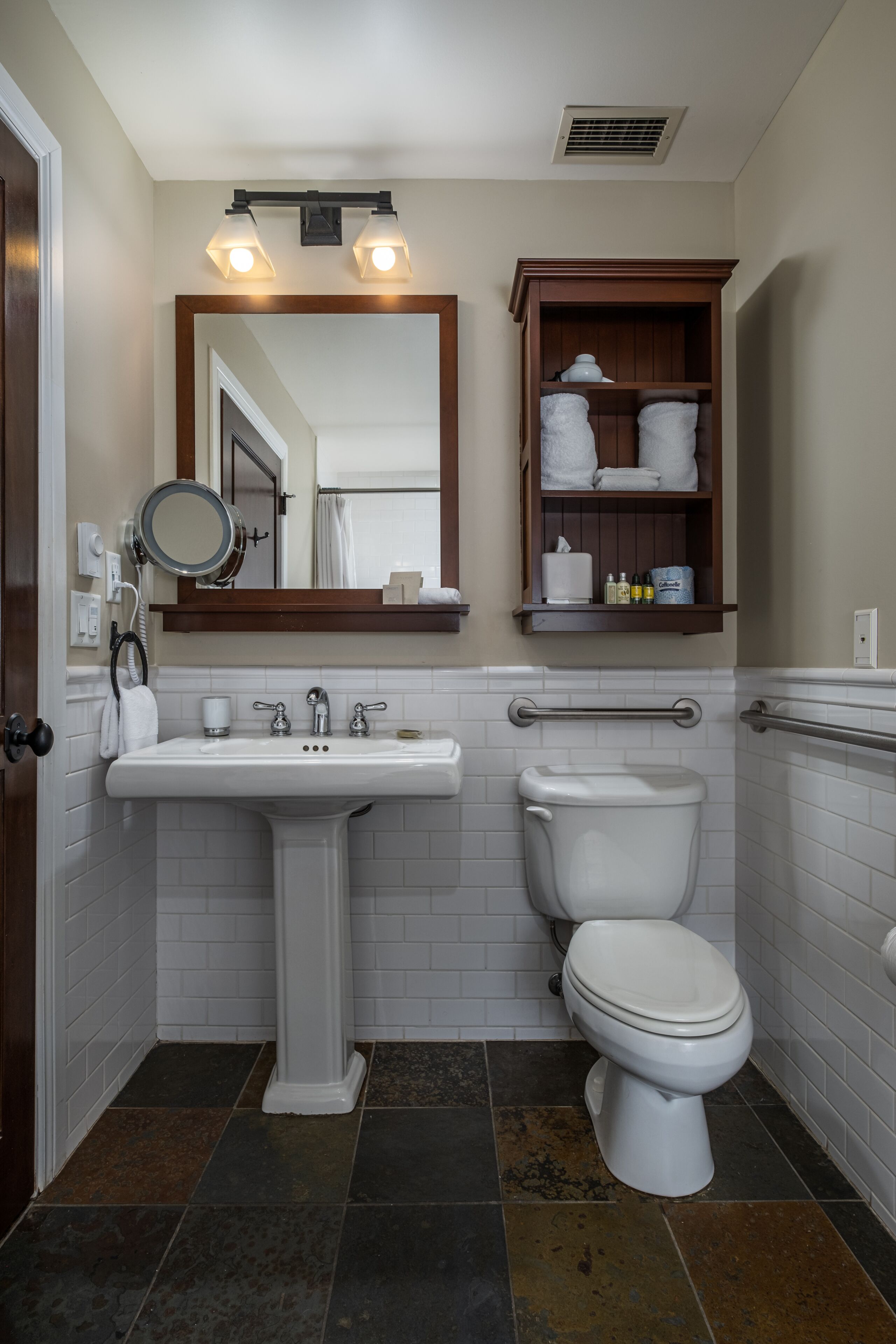 Un baño con lavamanos pedestal, inodoro, espejo y repisa con artículos de aseo.