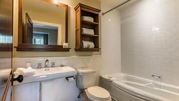 Un bany amb un lavabo blanc, vàter i banyera, prestatges de fusta sobre el vàter, i un mirall sobre el lavabo.
