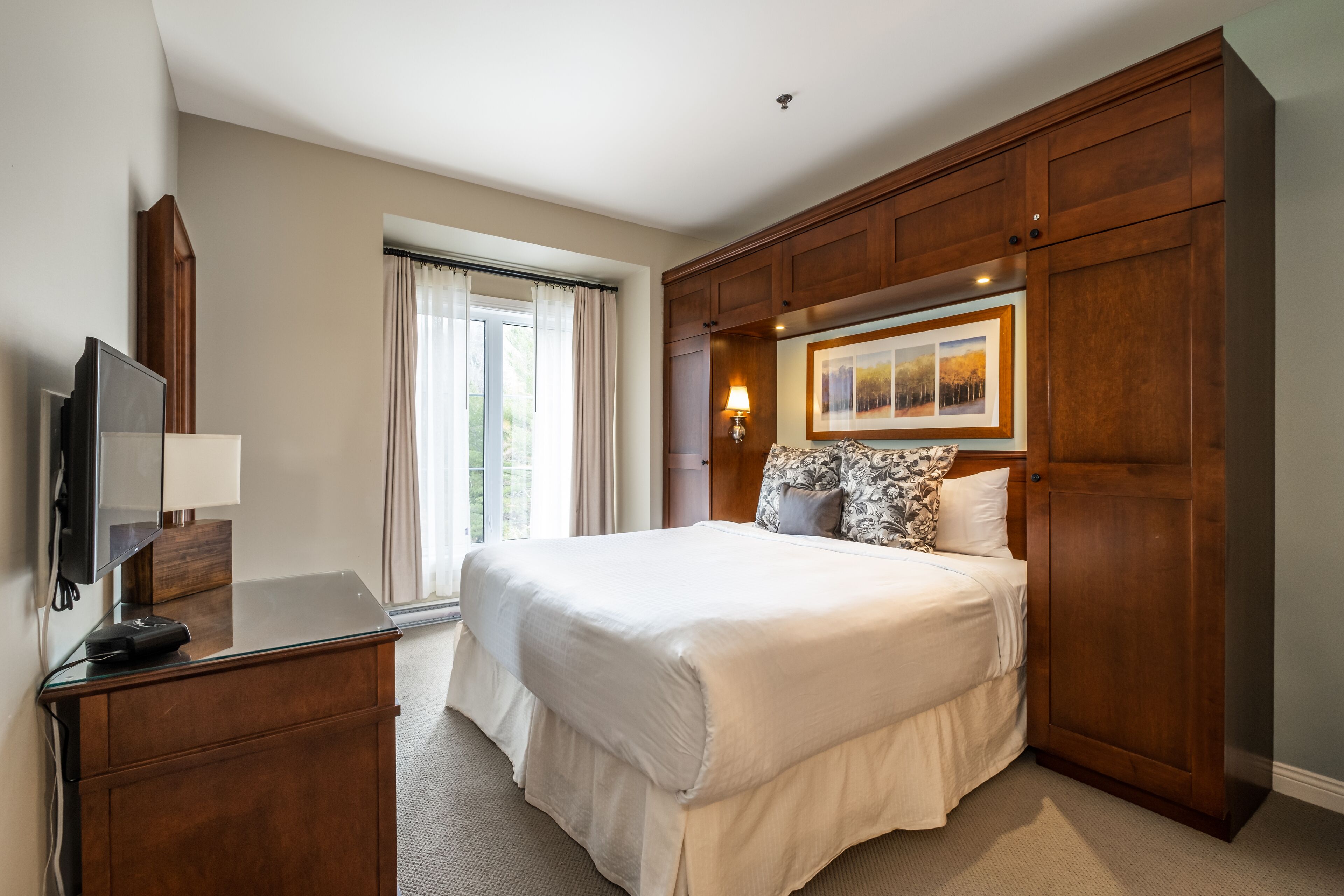 A hotel room with a large bed, wooden wardrobe, a desk with a flat-screen TV, and a window with curtains.