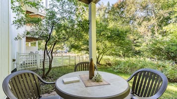 A round table with two chairs, a bottle, and a book on a patio with a white fence and trees in the background.