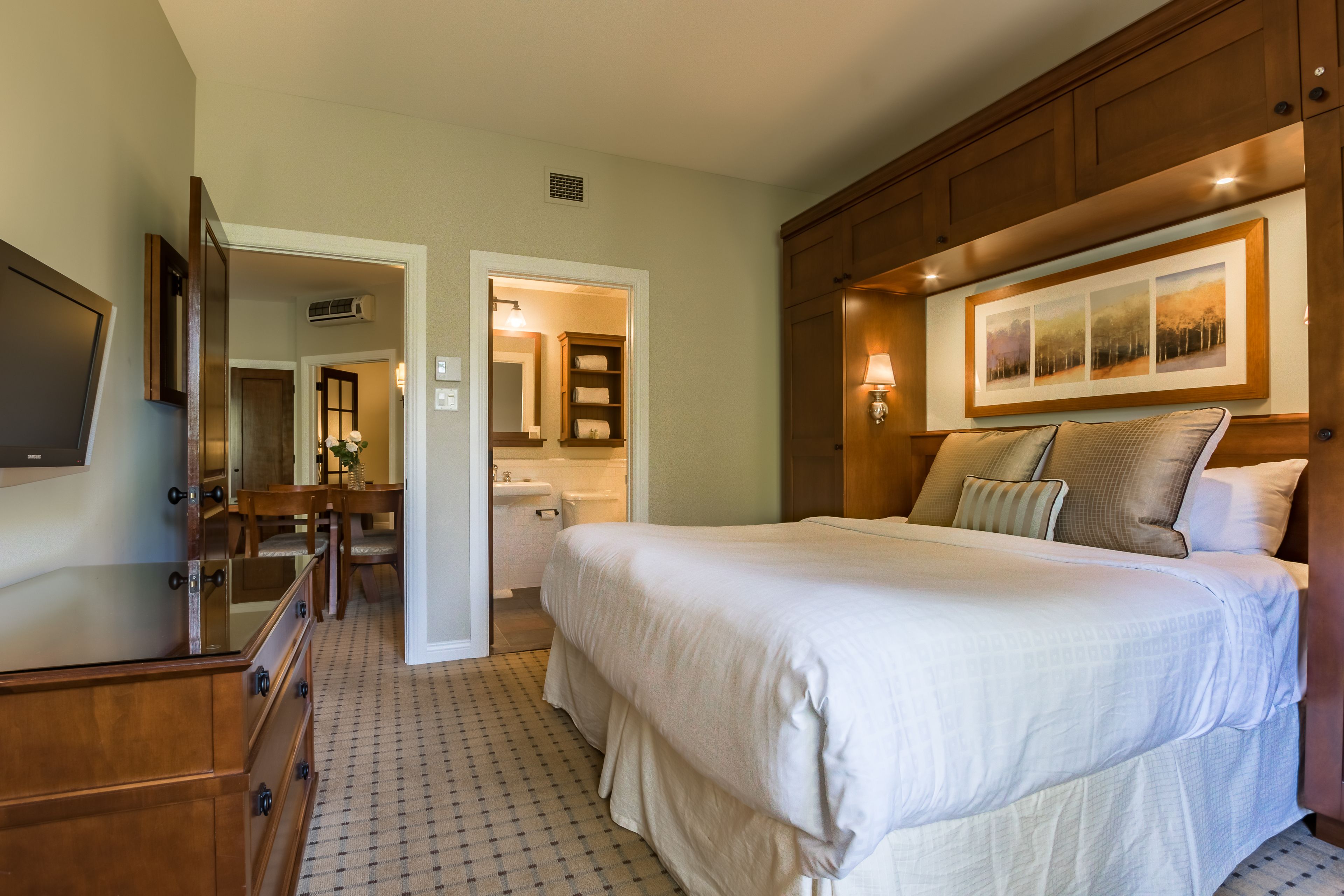 A hotel room with a large bed, wooden headboard, a flat-screen TV, a wooden dresser, and a bathroom visible through an open door.