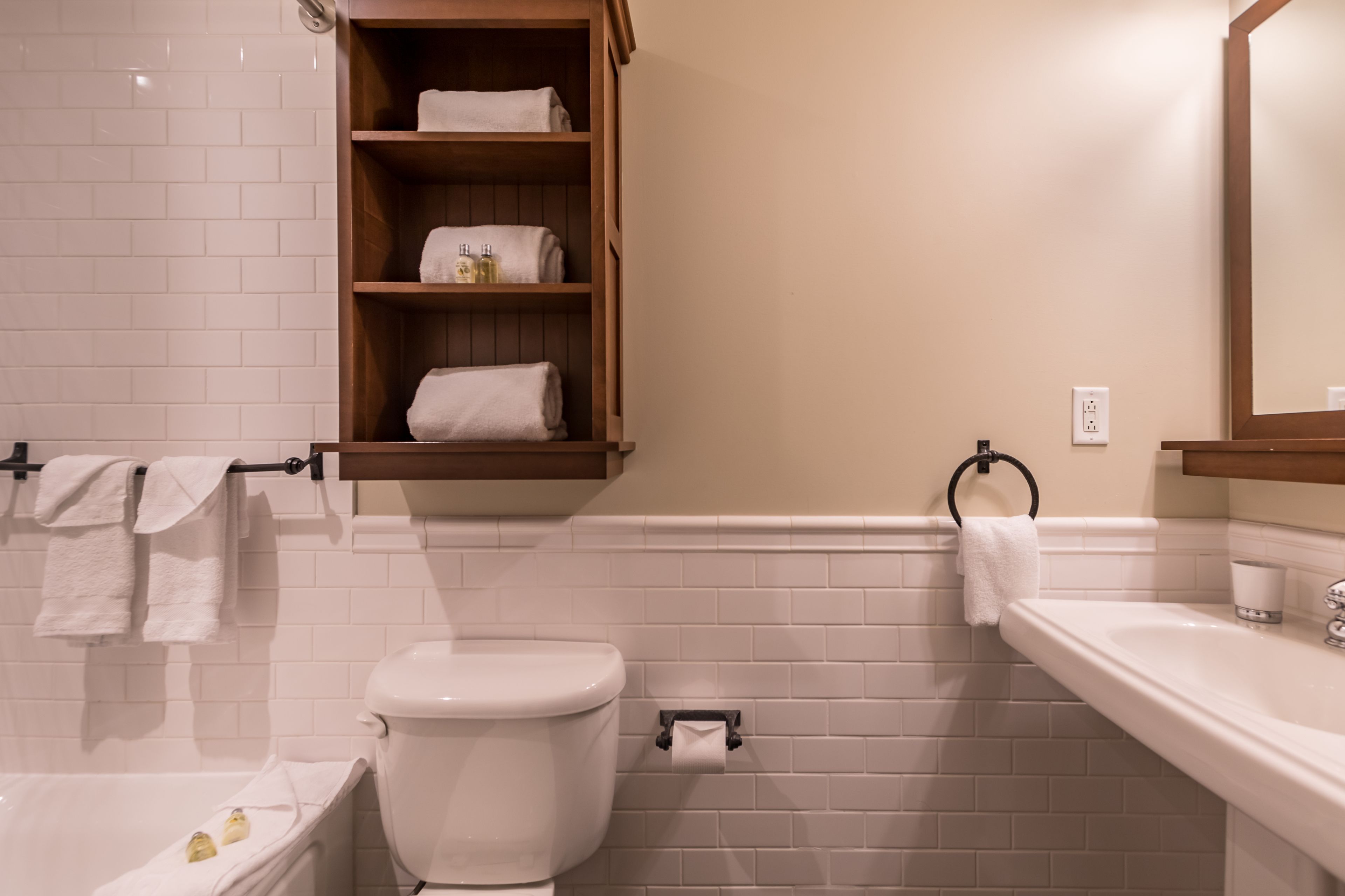 A bathroom with a toilet, sink, and bathtub. There are towels hanging on a rack and a shelf with toiletries.