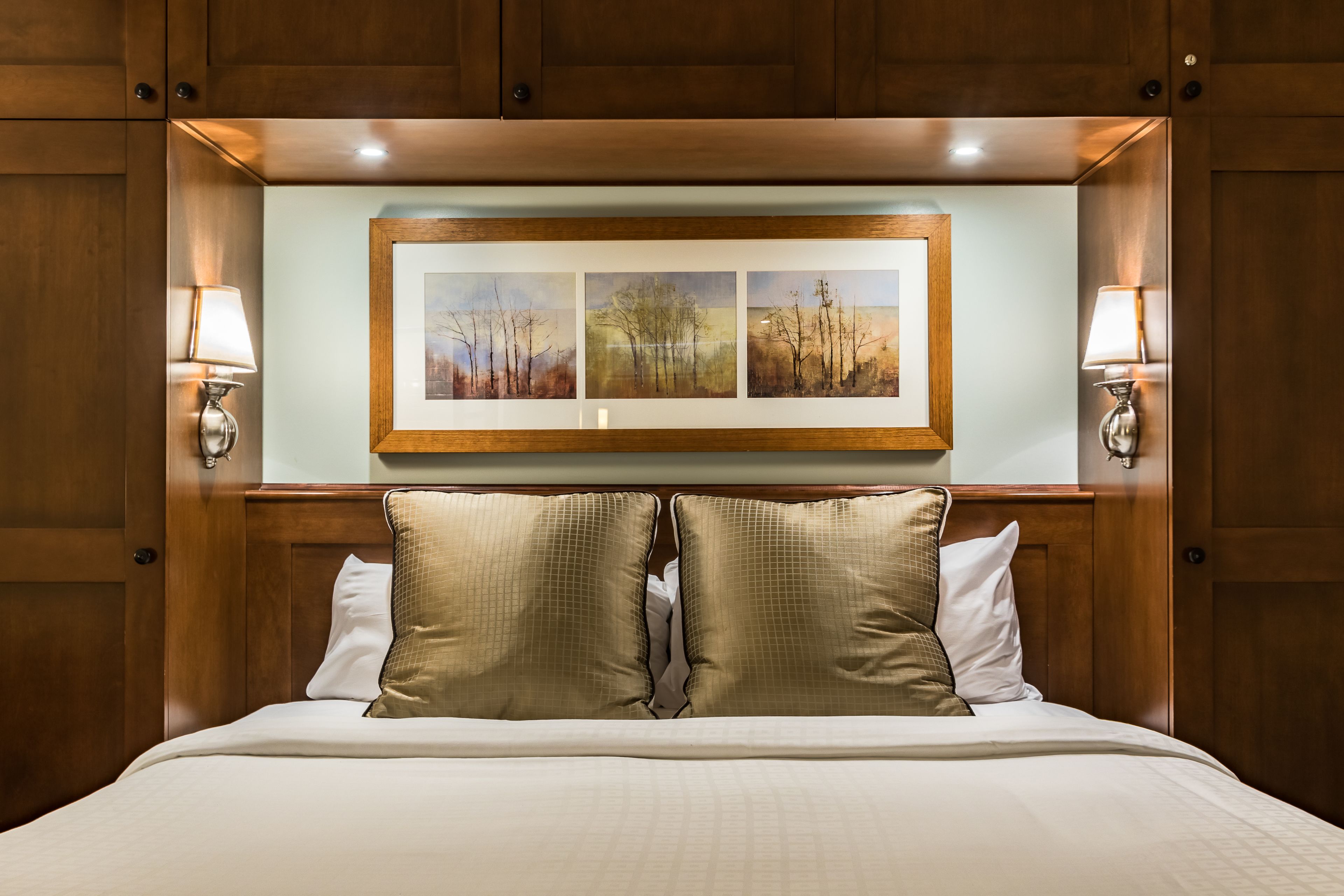 A hotel room with a wooden headboard, a framed landscape painting, two bedside lamps, and a bed with beige bedding.
