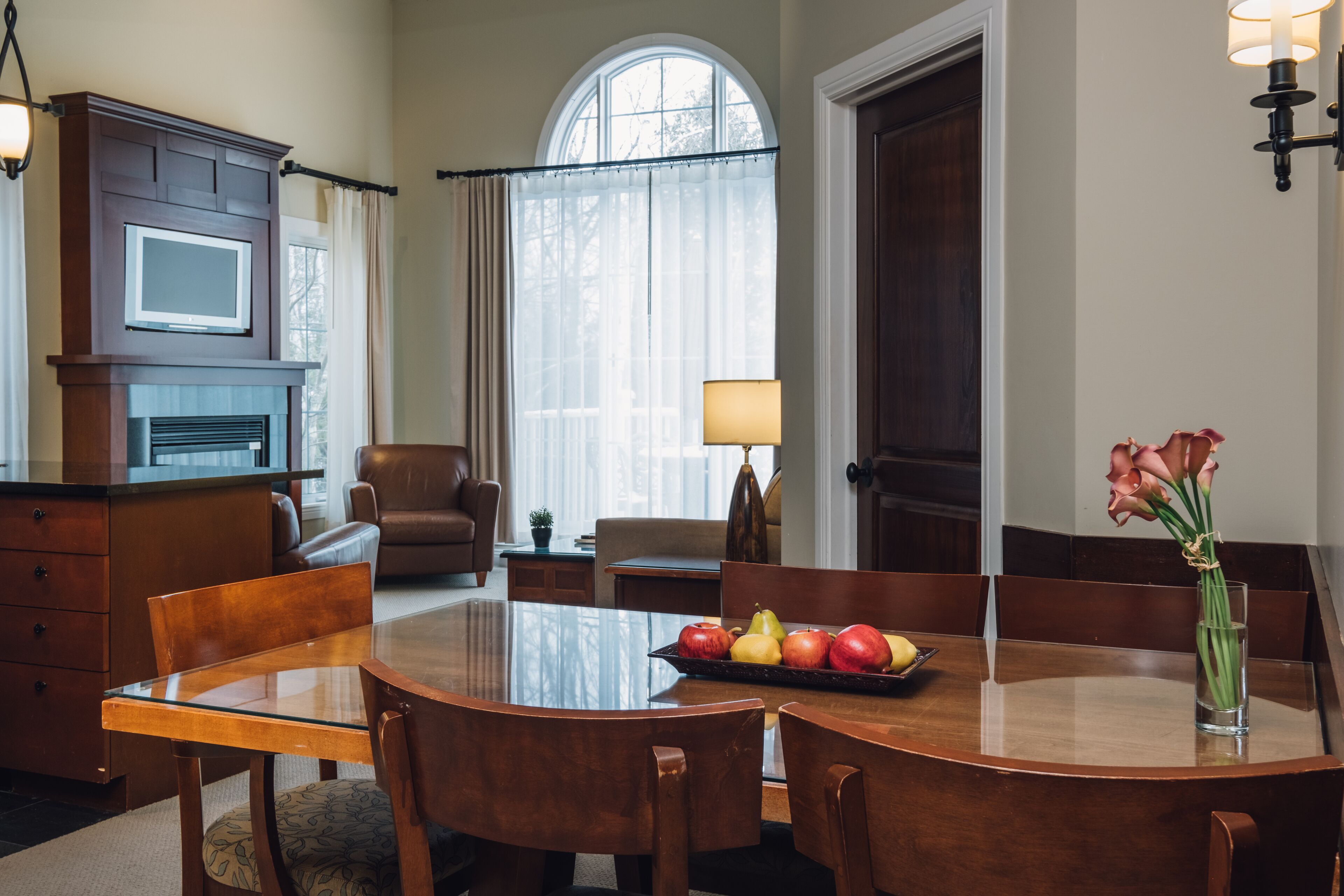 A dining area with a round table, chairs, and a fruit tray. There is a fireplace, a lamp, and a window with curtains.