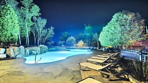Zona de piscina iluminada por la noche con sillones de descanso y árboles.