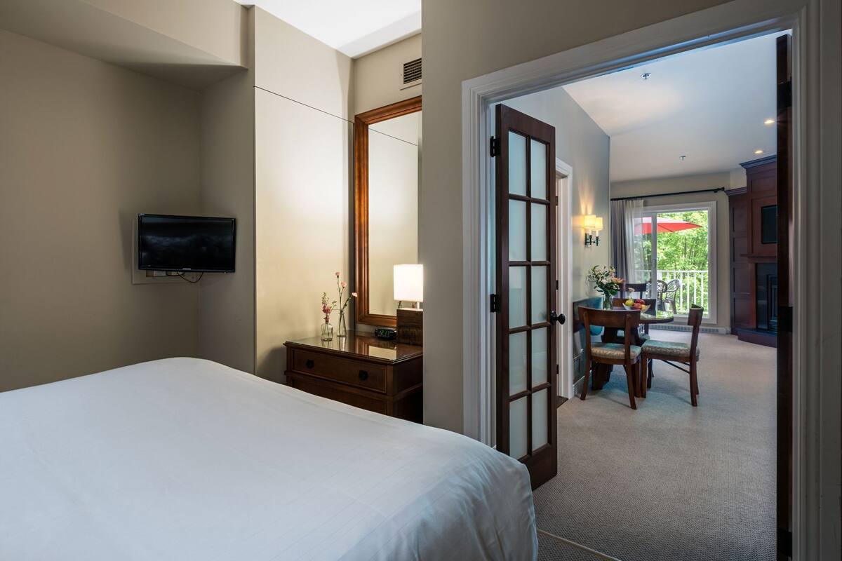 A hotel room with a bed, a TV, a dresser, a mirror, and a door leading to a balcony with outdoor seating.