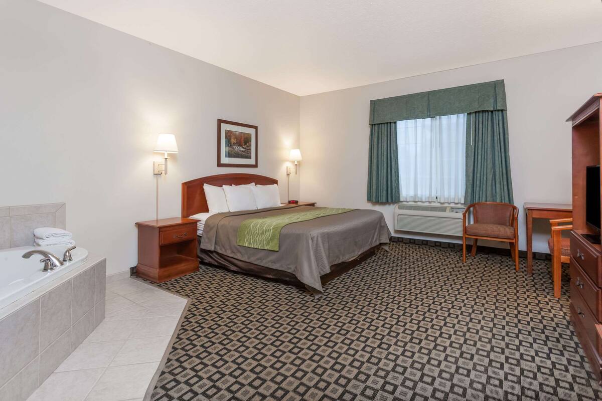 A hotel room with a bed, a desk, a chair, a desk lamp, a nightstand, a lamp, a window with curtains, a bathroom with a bathtub, and a shower.