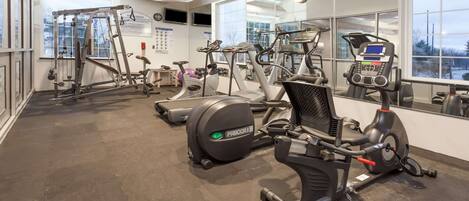 Ein Fitnessstudio mit verschiedenen Trainingsgeräten, großen Fenstern und einer Spiegelwand.