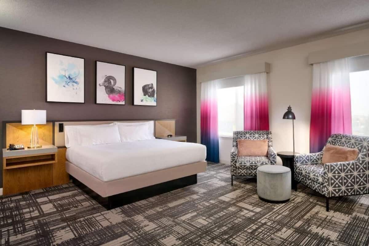A hotel room with a bed, two armchairs, a side table, a lamp, and artwork on the wall.