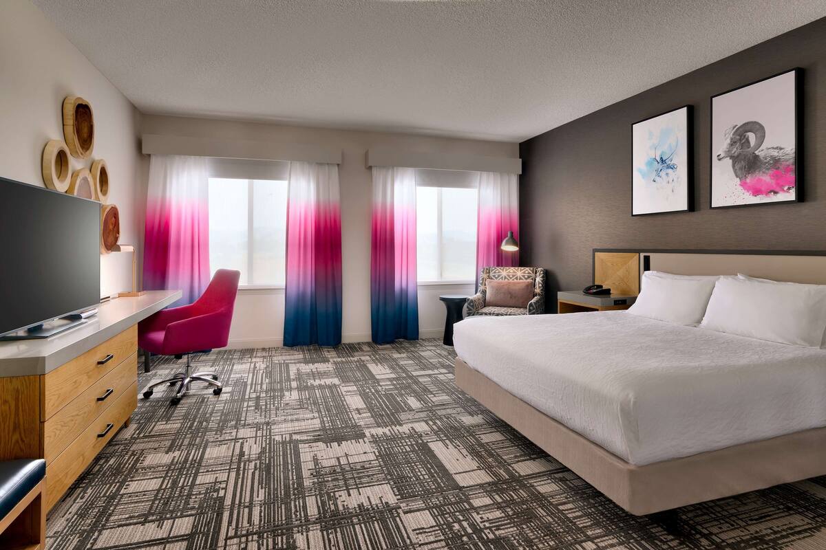 A hotel room with a large bed, a desk with a flat-screen TV, a chair, a small table, and a nightstand.