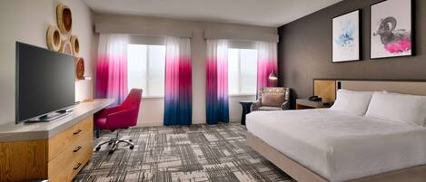 A hotel room with a large bed, a desk with a flat-screen TV, a chair, a small table, and a nightstand.