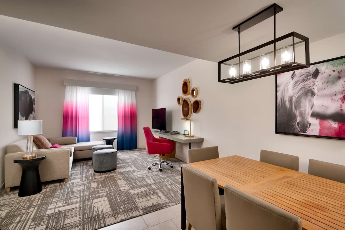 A modern hotel room with a wooden dining table, a sofa, a red chair, a TV, and a desk with a lamp.