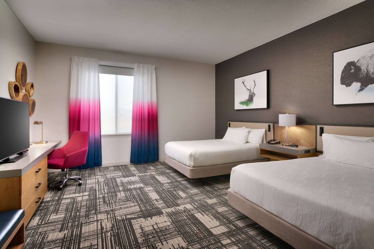 A hotel room with a TV, a desk with a lamp, a chair, two beds, a bedside table with a lamp, and artwork on the walls.