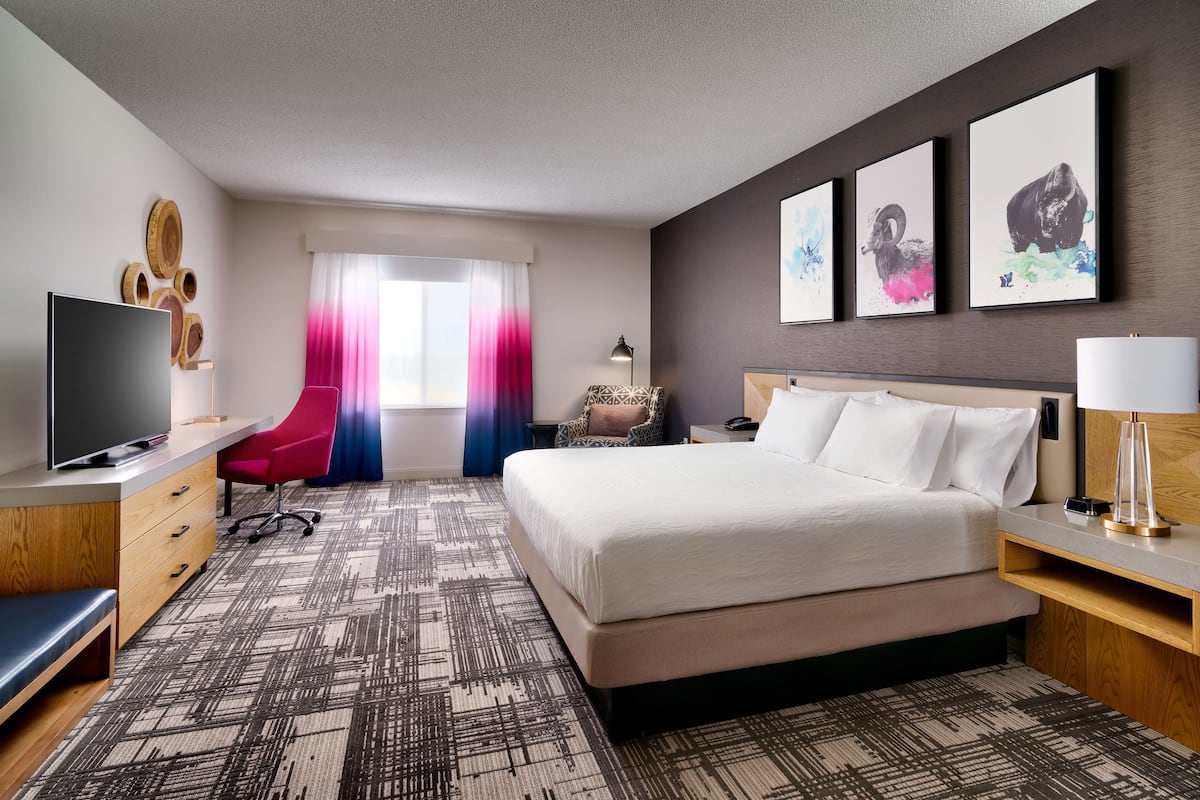 A hotel room with a bed, a desk with a computer, a chair, a TV, and artwork on the wall.