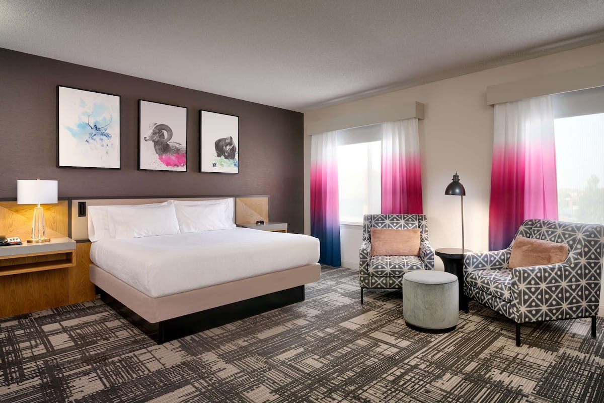 A hotel room with a bed, two armchairs, a side table, a lamp, and three wall-mounted artworks.