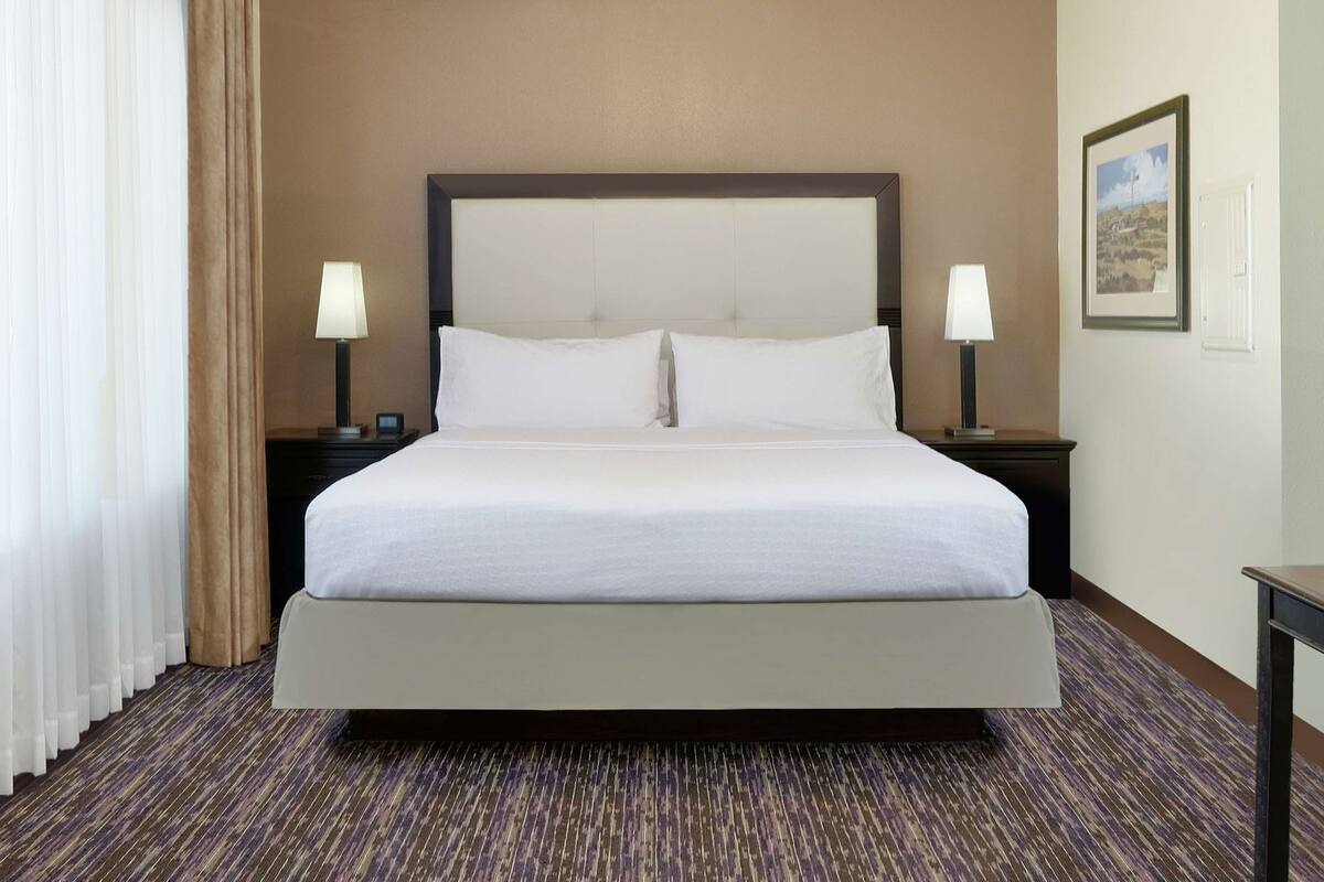 A hotel room with a large bed, two bedside tables with lamps, a carpeted floor, and a framed picture on the wall.