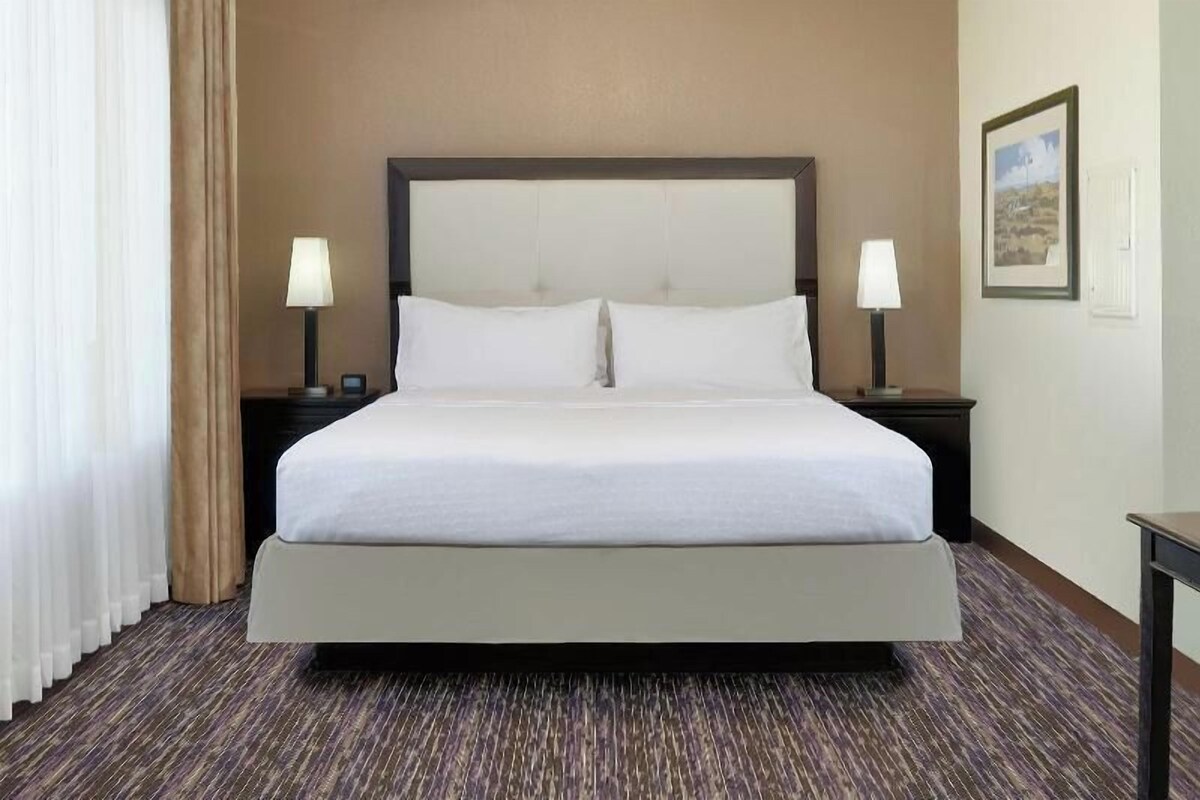 A hotel room with a large bed, two bedside tables with lamps, a framed picture on the wall, and a carpeted floor.