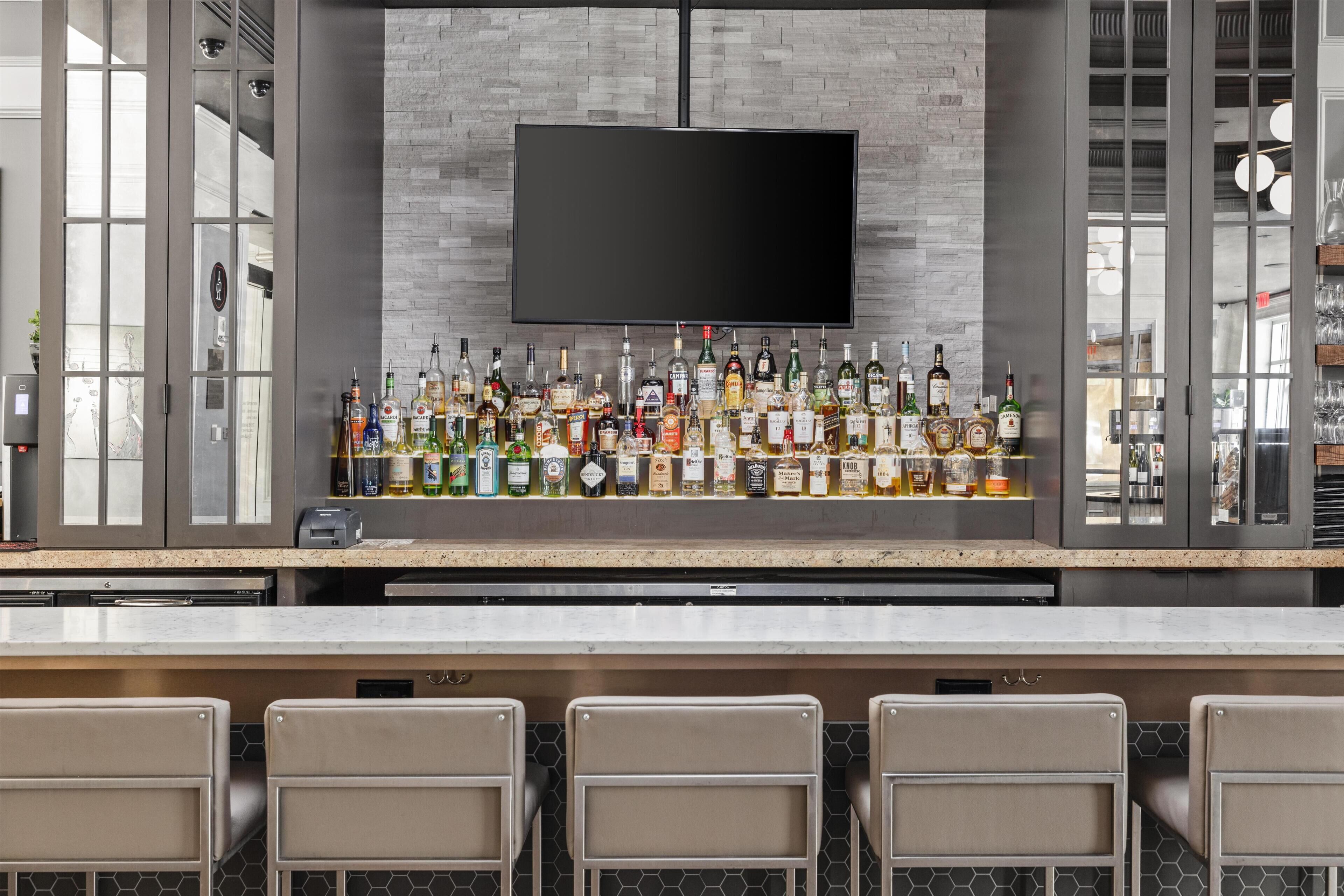 A bar area with a flat-screen TV mounted on a brick wall, a variety of liquor bottles on the counter, and four bar stools.