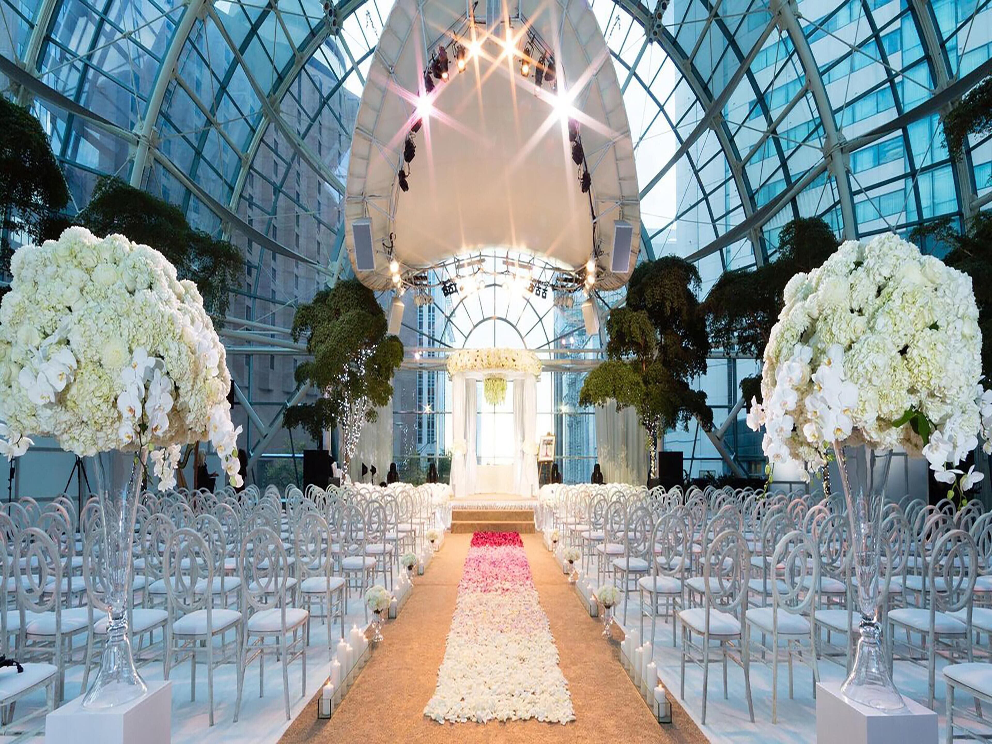 A grand hall with a glass ceiling, white chairs arranged in rows, and large white floral arrangements.