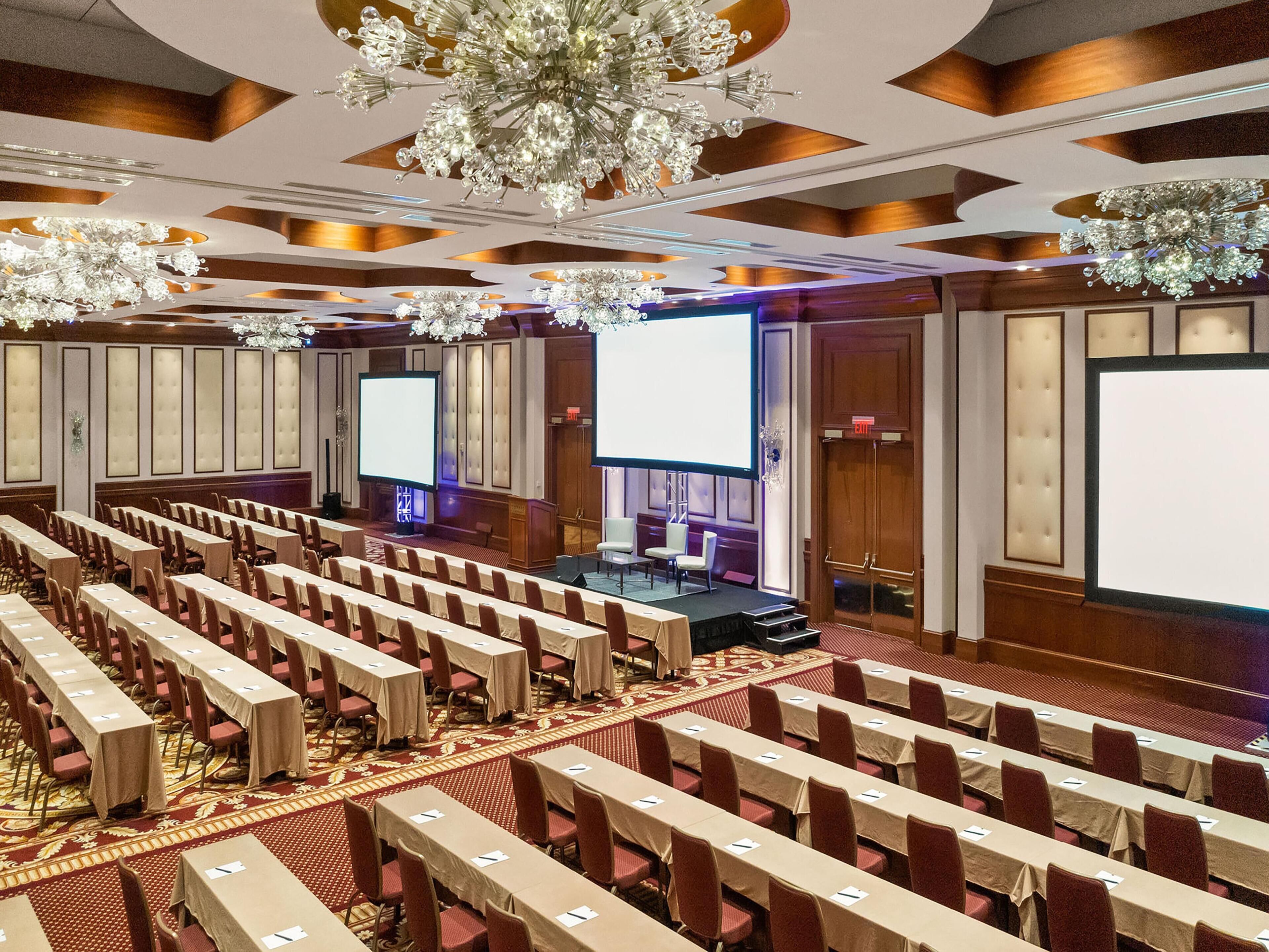 A conference room with rows of tables, a stage, and large screens.