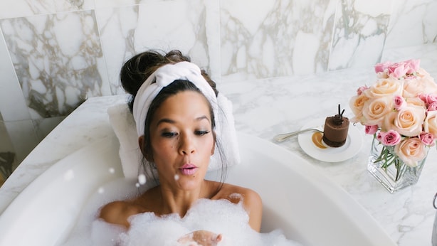 A person in a bathtub filled with bubbles, holding a glass of champagne, with a cake and flowers on the side. - Conrad Indianapolis (Indianapolis)