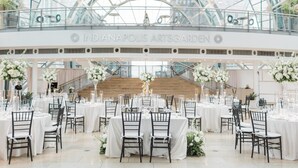 A spacious indoor venue with multiple round tables and chairs, a glass ceiling, and a central staircase. - Conrad Indianapolis (Indianapolis)
