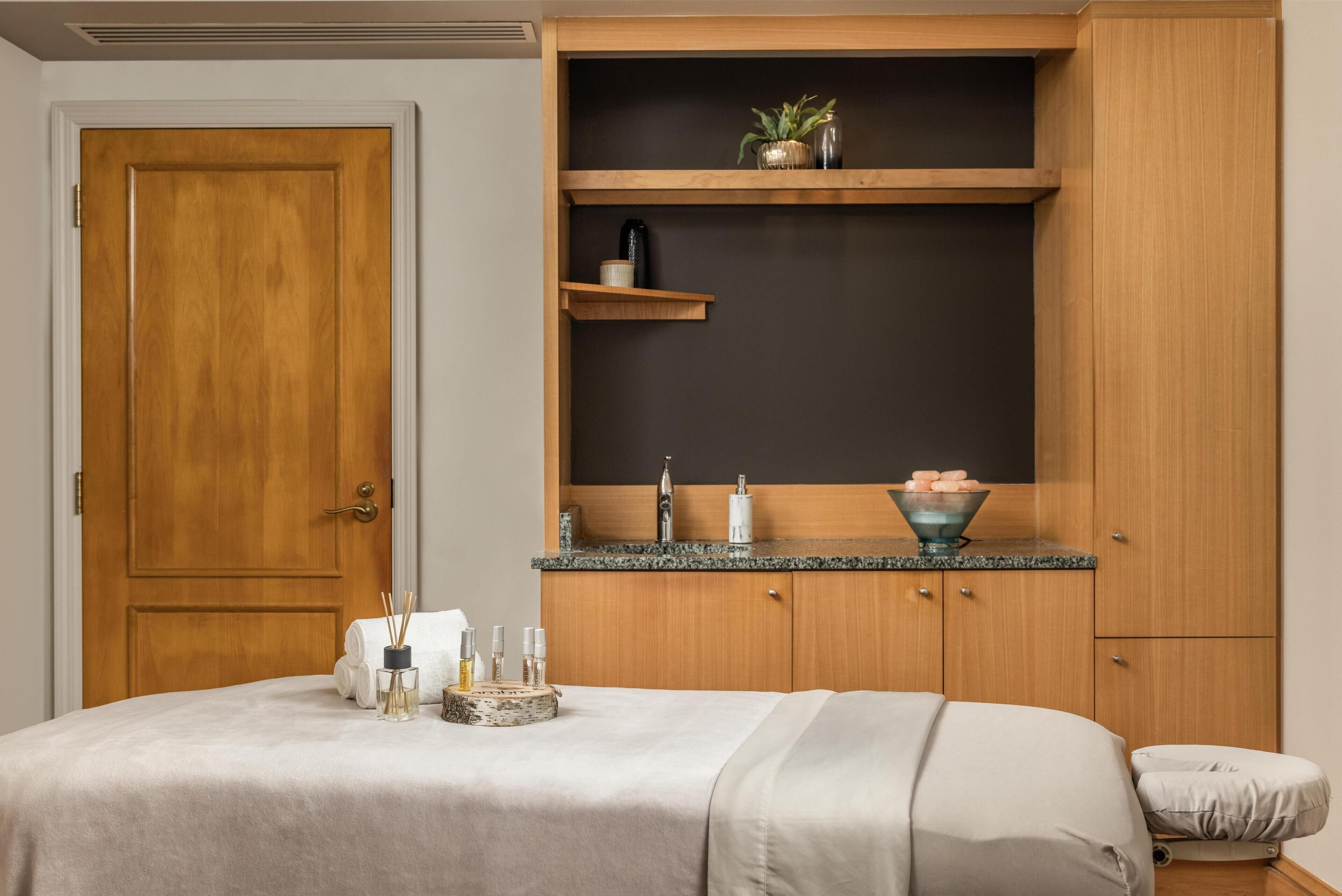 A bedroom with a wooden door, a bed with a beige bedspread, a small bedside table with toiletries, a shelf with decorative items, and a dark wall behind the bed.