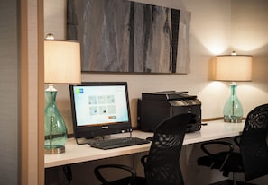 A hotel room with a desk, a computer, a printer, two lamps, and a decorative wall piece.