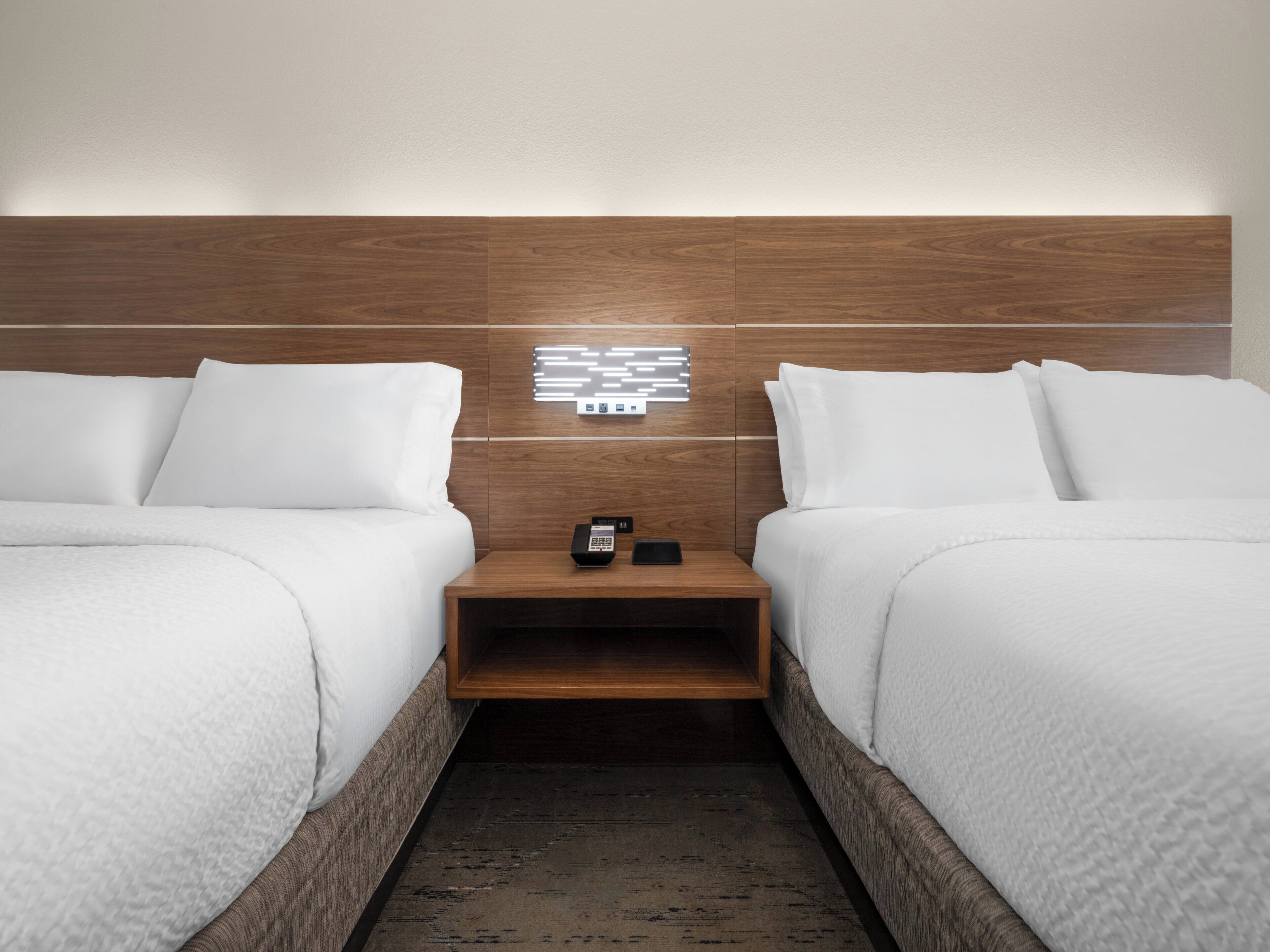 Habitación de hotel con dos camas, cabecera de madera, mesita de noche con teléfono y lámpara fijada a la pared.