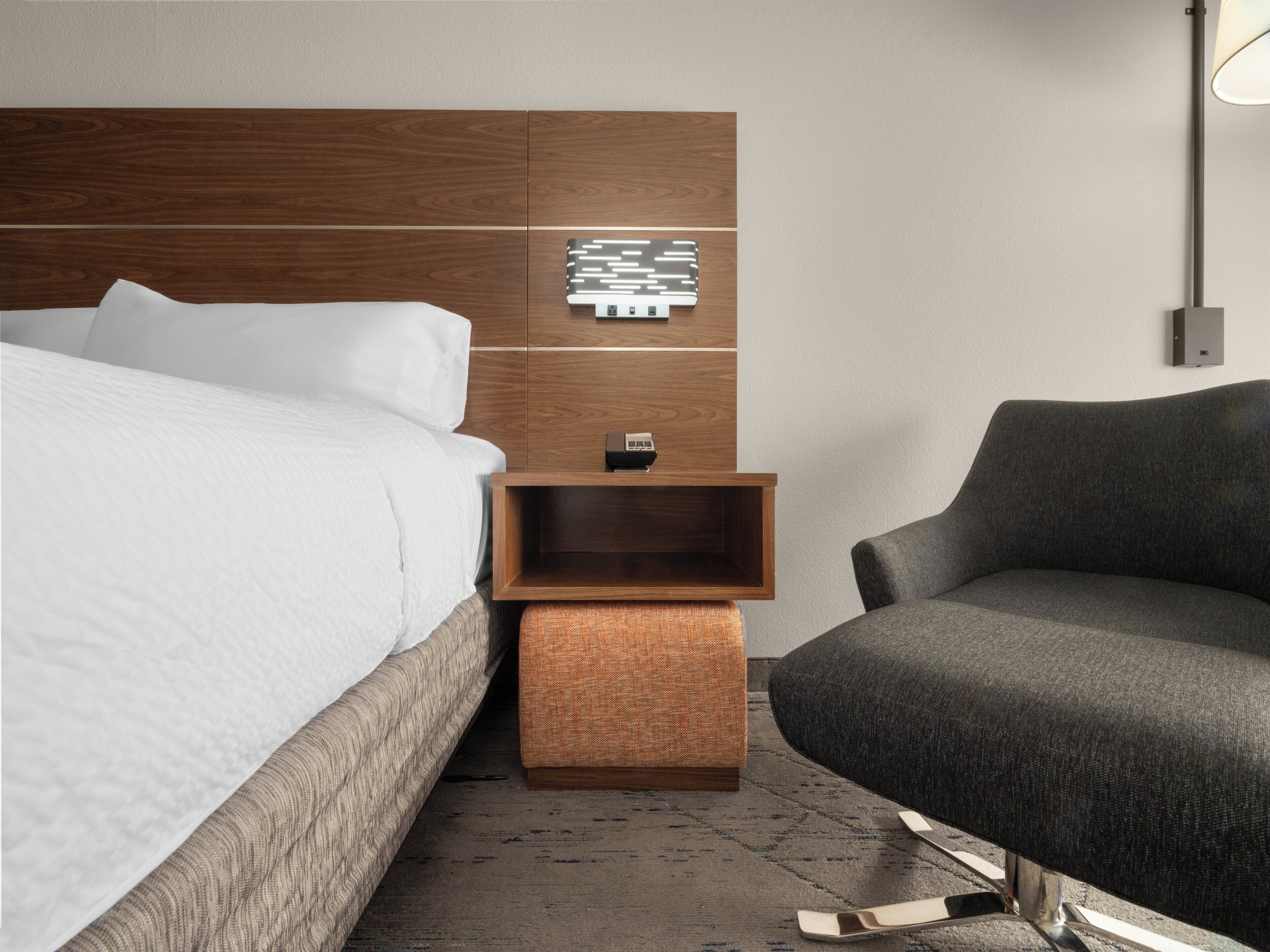 Une chambre d’hôtel moderne avec une tête de lit en bois, une table de chevet avec une lampe, un fauteuil gris et un pouf à motifs.