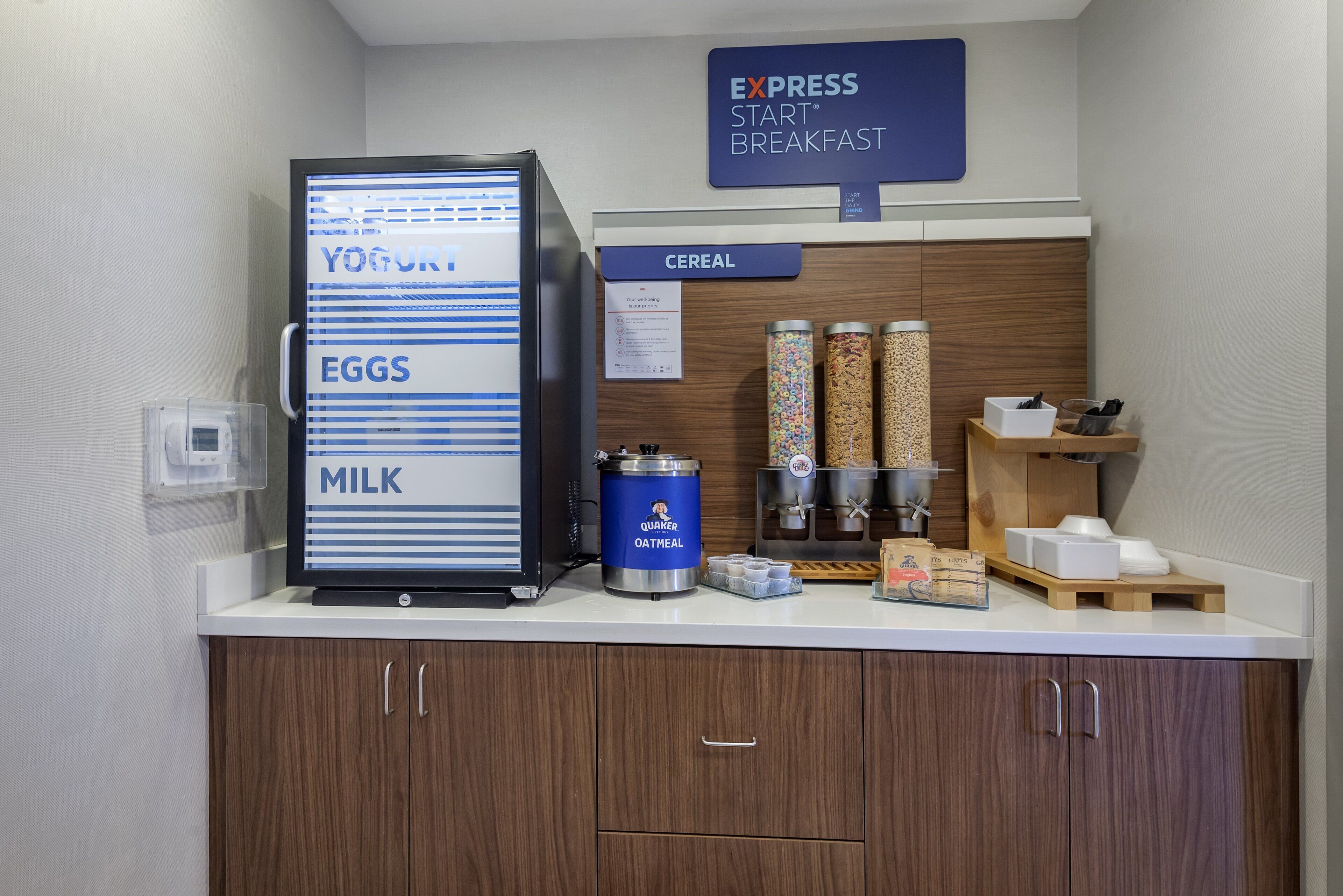 A breakfast station with a refrigerator displaying yogurt, eggs, and milk, a Quaker Oatmeal container, and cereal dispensers.