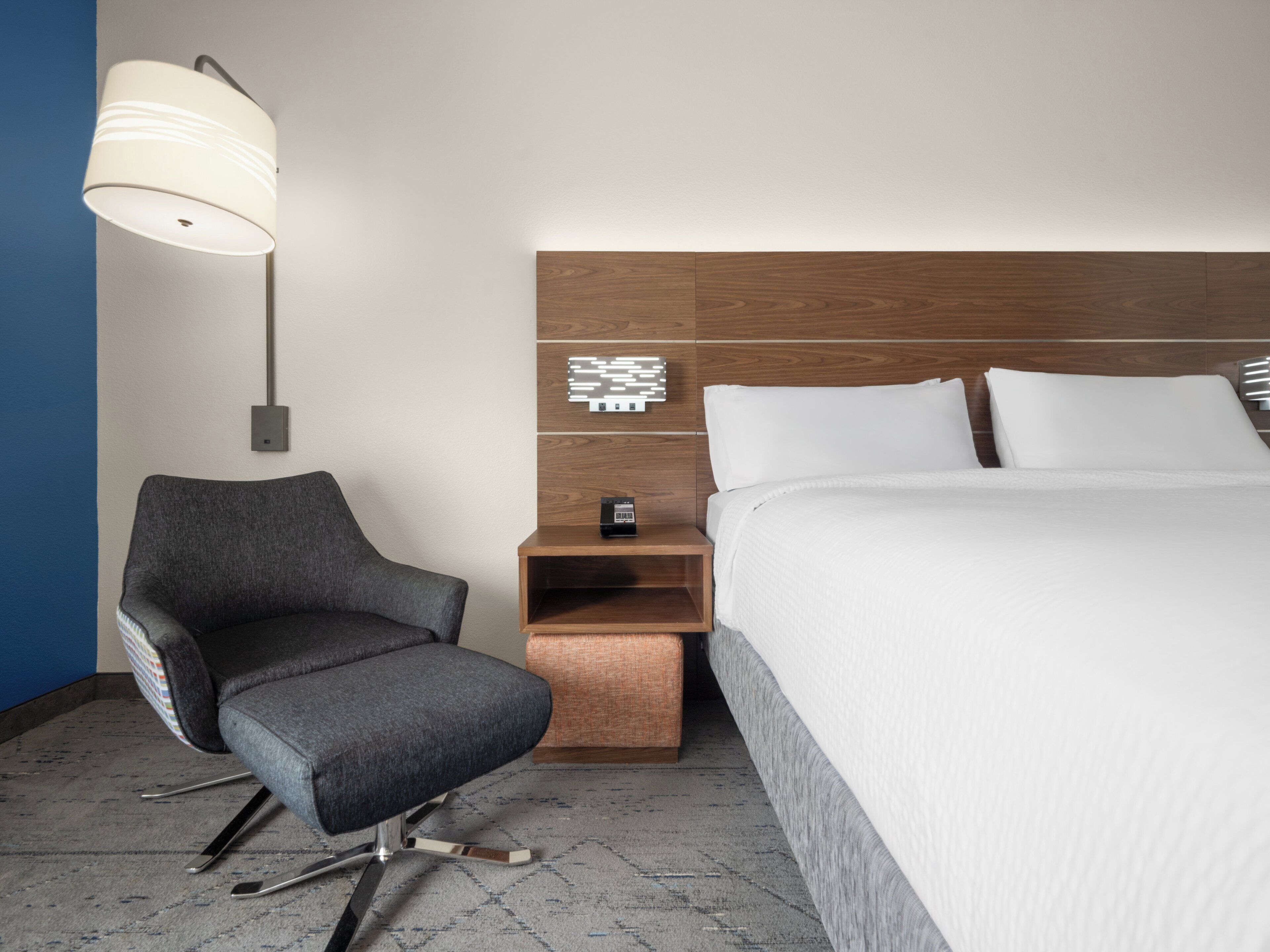 Habitación de hotel con cama, una silla, mesita de noche, lámpara y panel de control en la pared.