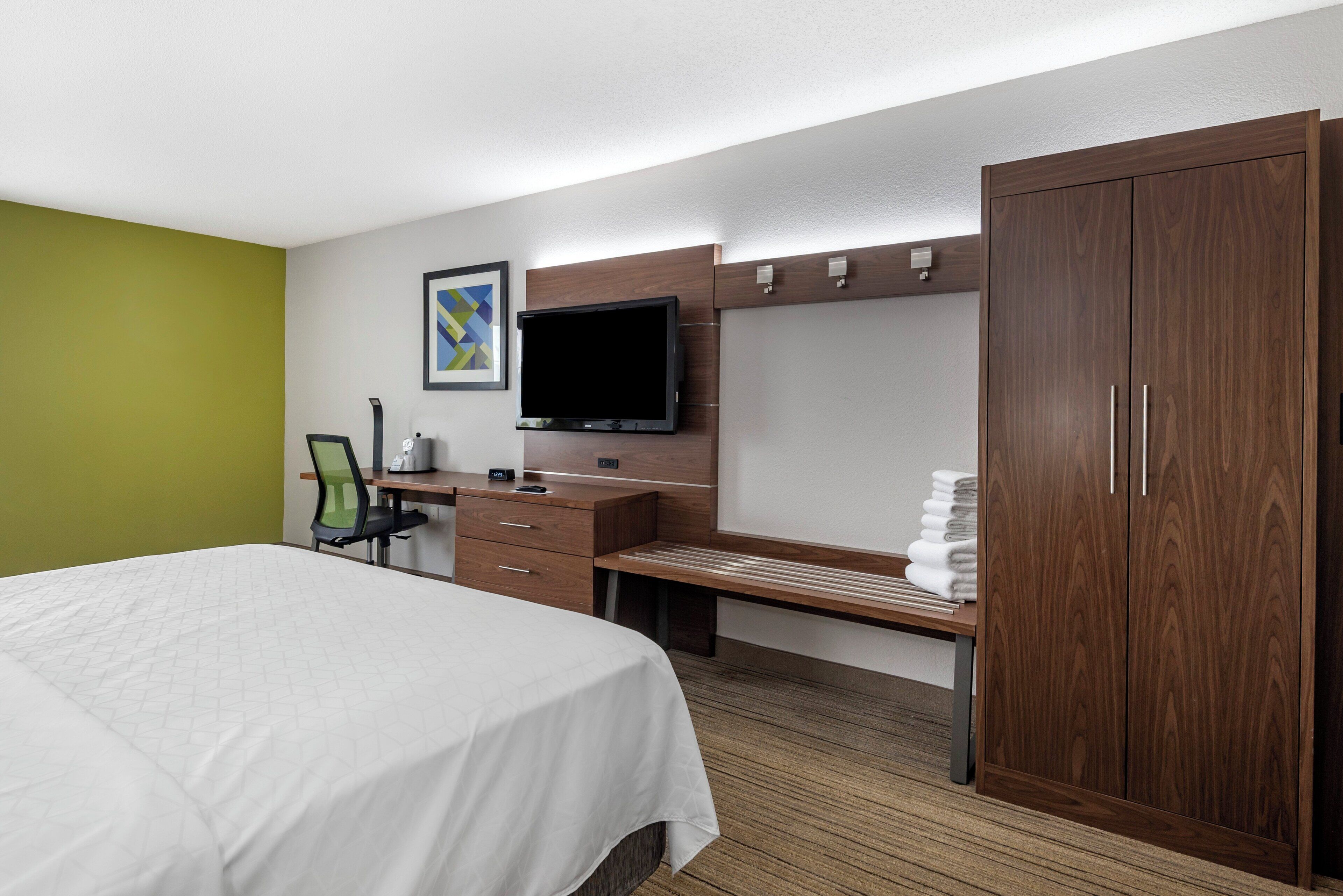 Habitación de hotel con cama, escritorio, silla, televisor, armario y un cuadro en la pared.