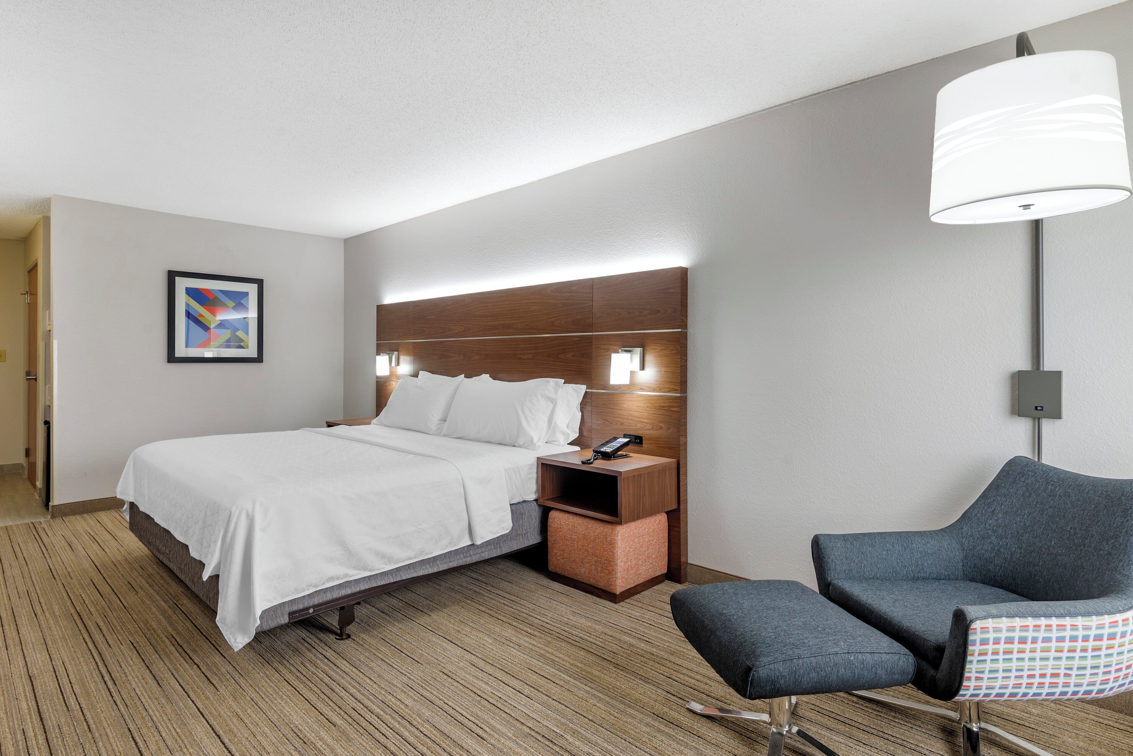 Una habitación de hotel con cama, mesita de noche, silla y un cuadro en la pared.