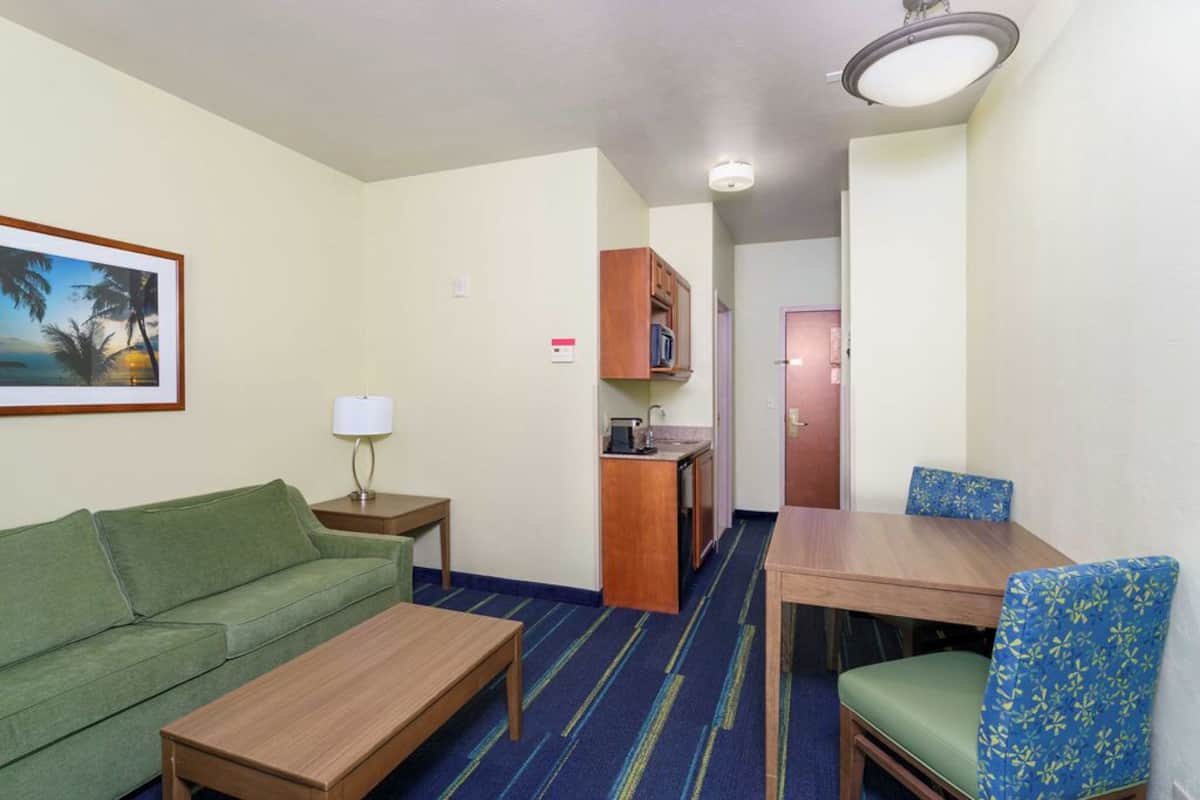 A compact hotel room with a green sofa, a wooden table, a blue-striped carpet, a kitchenette, and a door leading to another room.