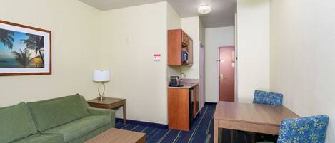 Une chambre d’hôtel compacte avec un canapé vert, une table en bois, un tapis à rayures bleues, une kitchenette et une porte donnant sur une autre pièce.