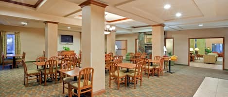 A spacious hotel dining area with multiple tables and chairs, a patterned carpet, and a television mounted on the wall.