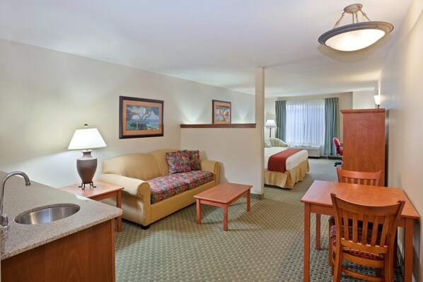 A hotel room with a kitchenette, a sofa, a small table, a wooden chair, a bed, and a window with curtains.