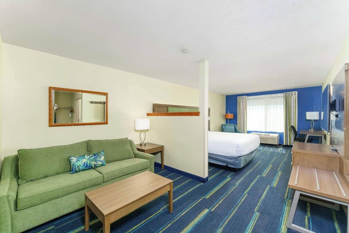 A hotel room with a green sofa, a wooden coffee table, a bed with a white comforter, a desk with a chair, a mirror, and a window with curtains.
