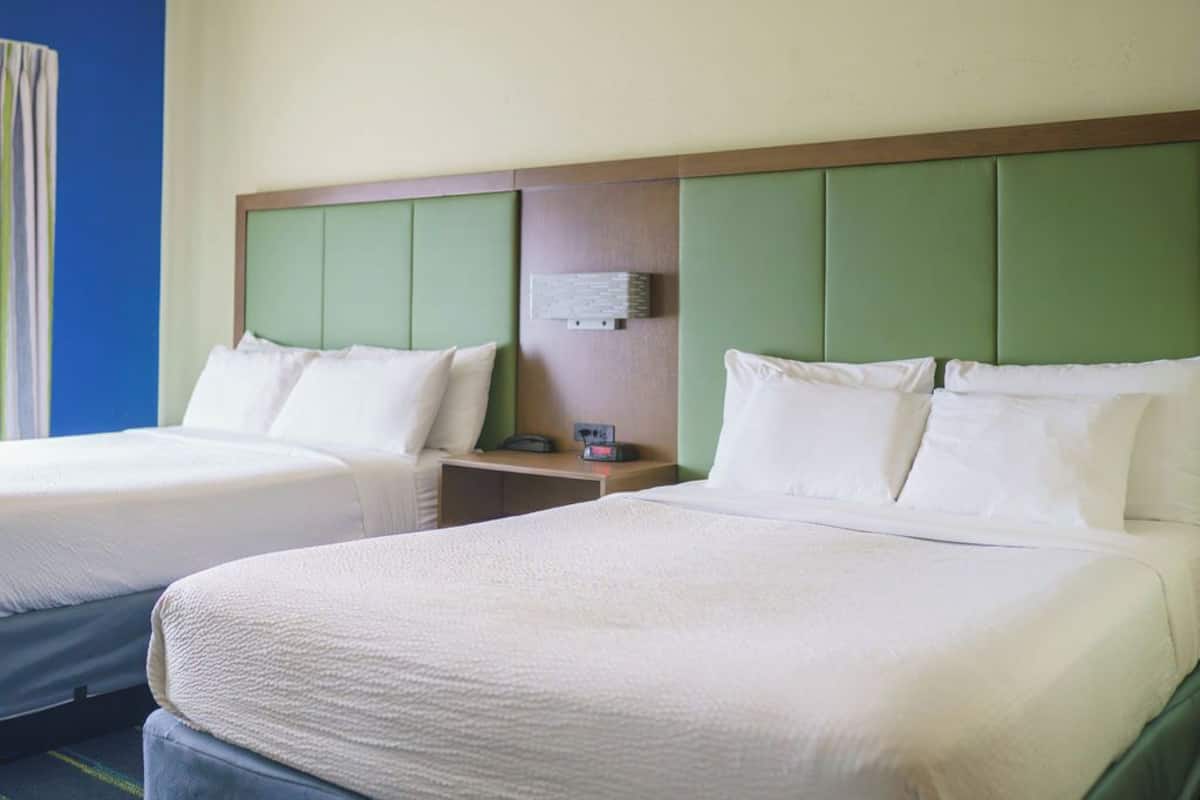 A hotel room with two beds, each with white linens and a green headboard. There is a small bedside table with a lamp and a power outlet.