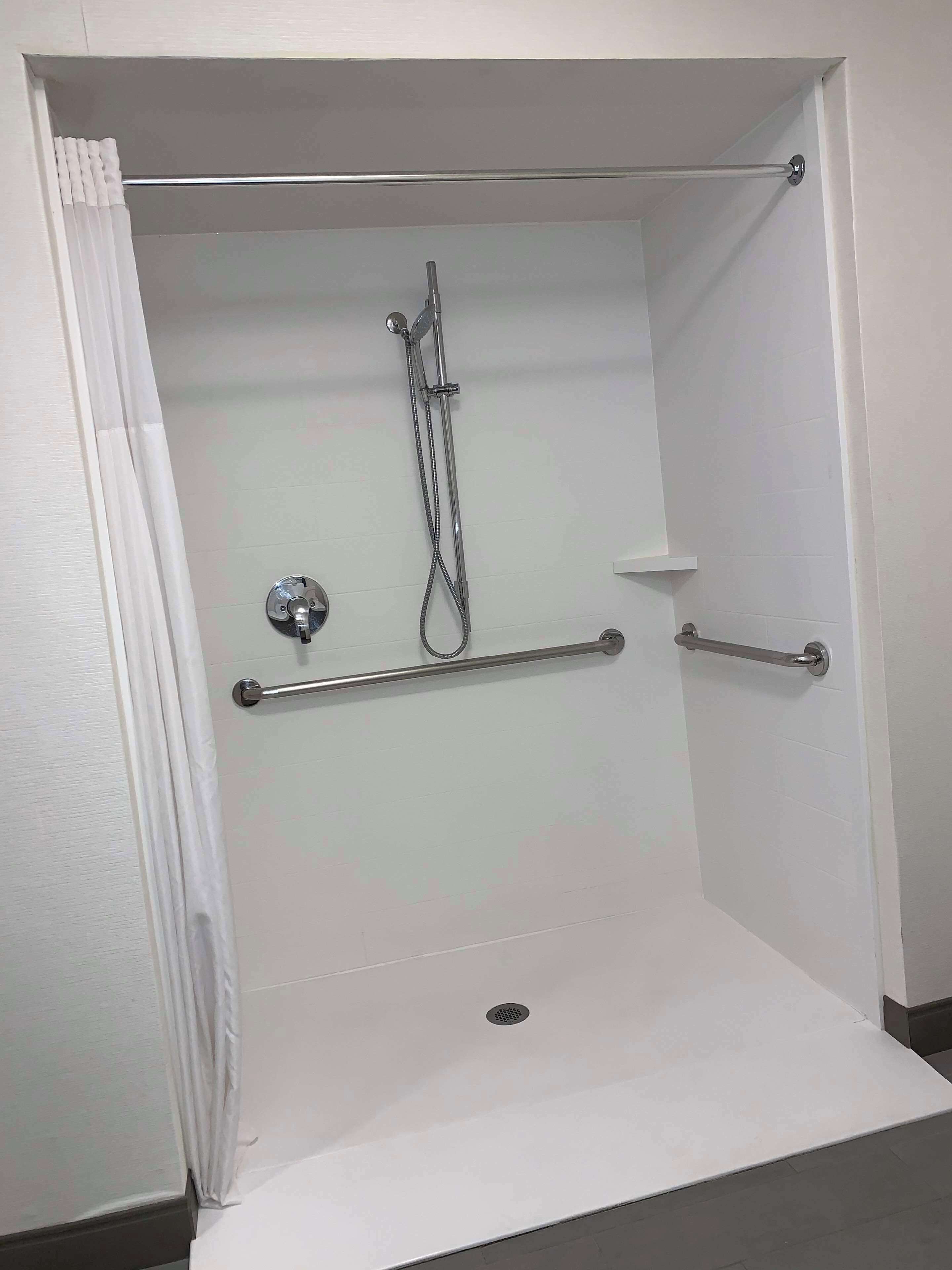 A shower area with a glass door, a handheld showerhead, a handheld shower attachment, a grab bar, and a small shelf.