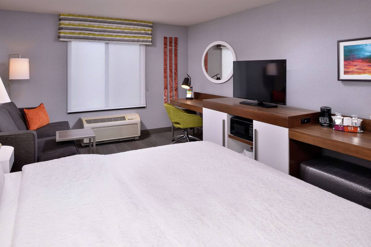 A hotel room with a bed, a desk with a computer, a TV, a chair, a lamp, and a window with curtains.