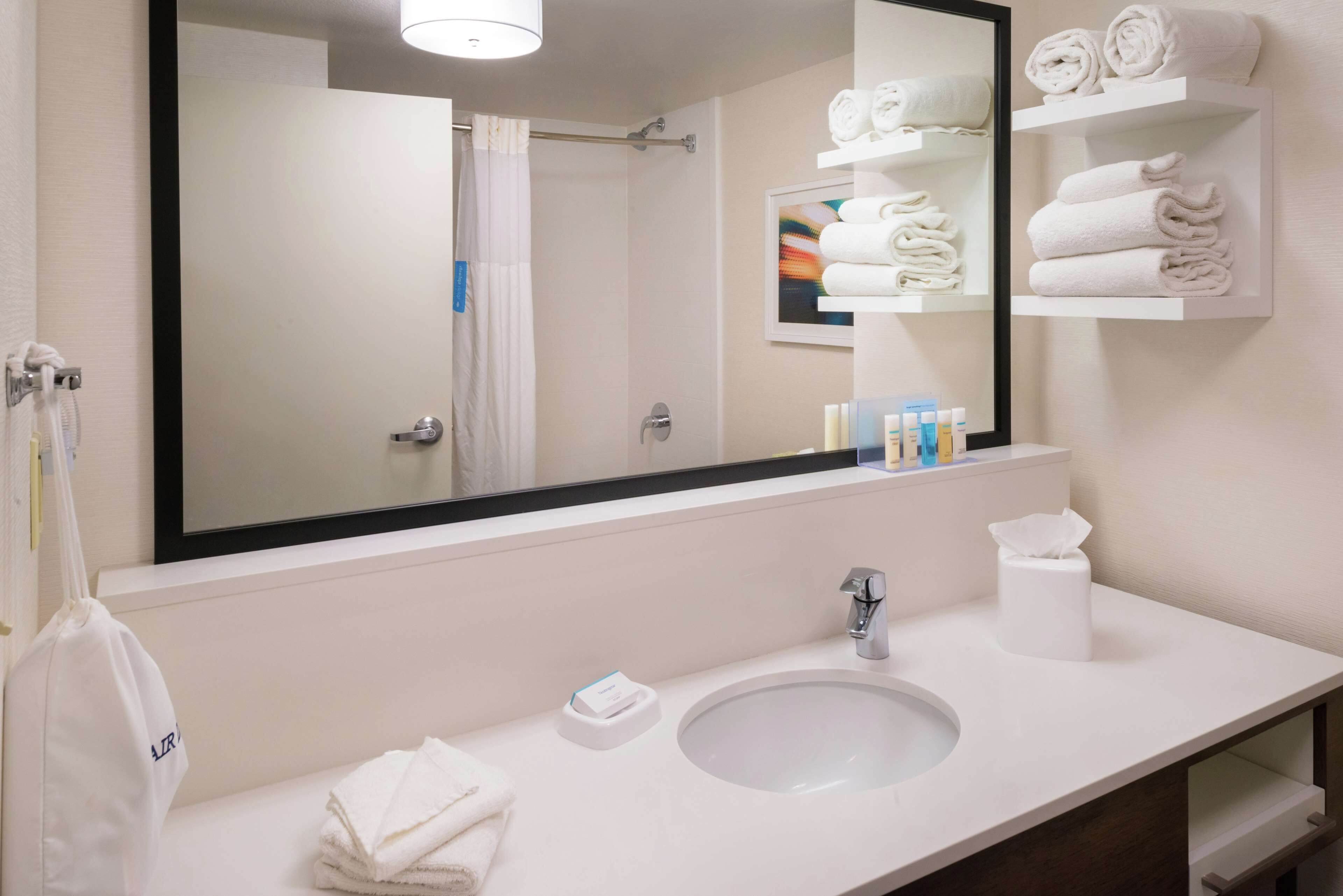 Un bany d'hotel amb un mirall gran, un lavabo blanc, un suport de tovalloles i prestatges amb tovalloles doblegades.