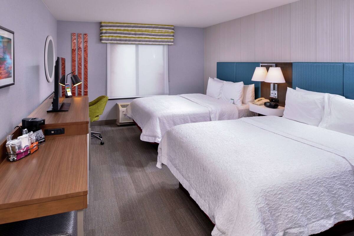 A hotel room with two beds, a desk, a chair, a mirror, a phone, and a window with blinds.